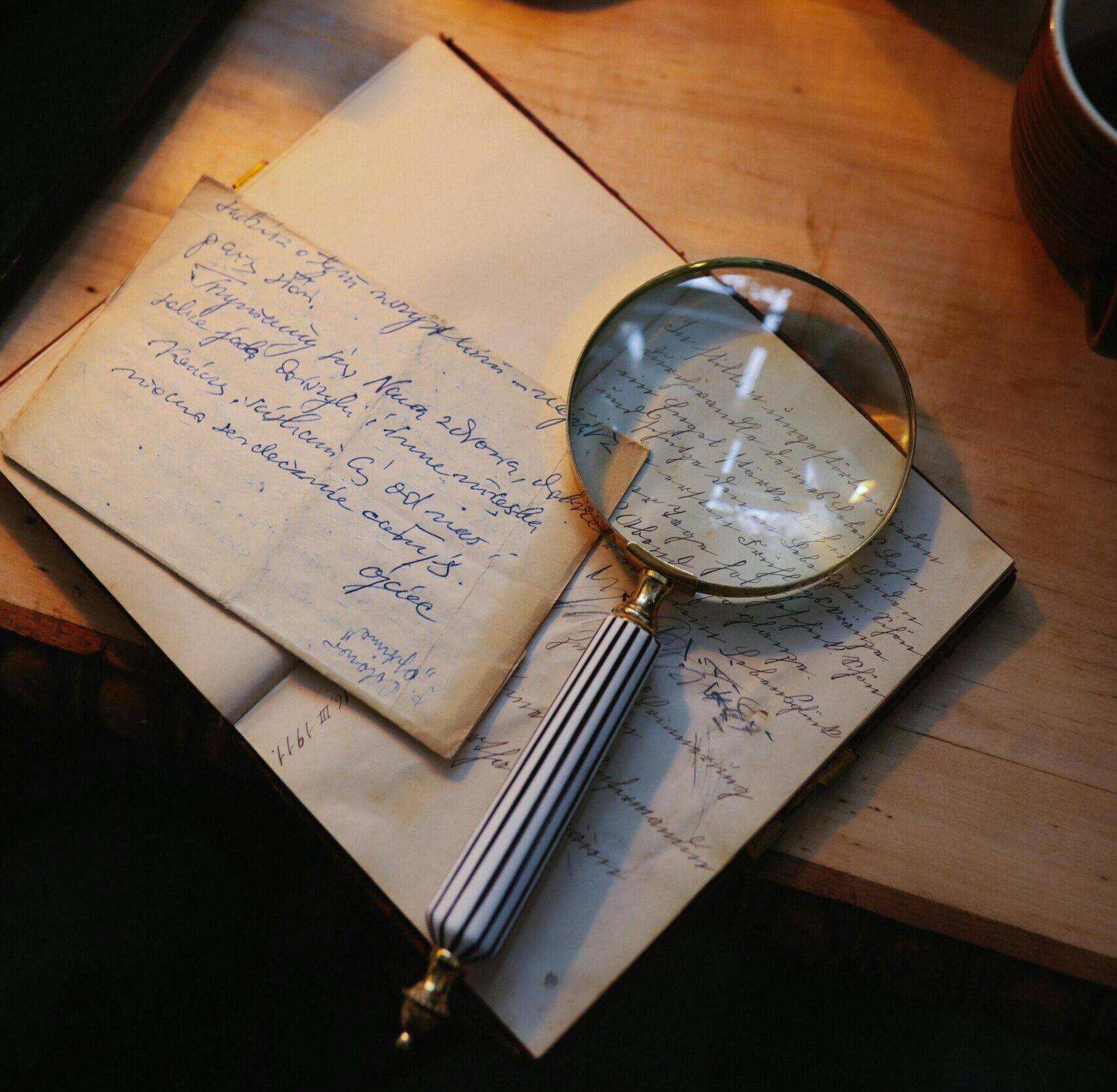 A brass magnifying glass resting on aged handwritten letters and documents on a wooden desk, with a vintage alarm clock in the background